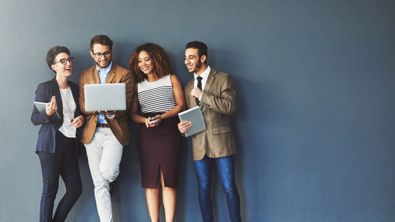 People Hero A group of four business people standing in front of a gray wall, looking at laptop and holding notebooks.